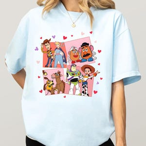 May include: Pale blue t-shirt with a vibrant graphic of Toy Story characters. The design showcases Woody, Buzz Lightyear, Jessie, and others, set against pink squares and red heart motifs.