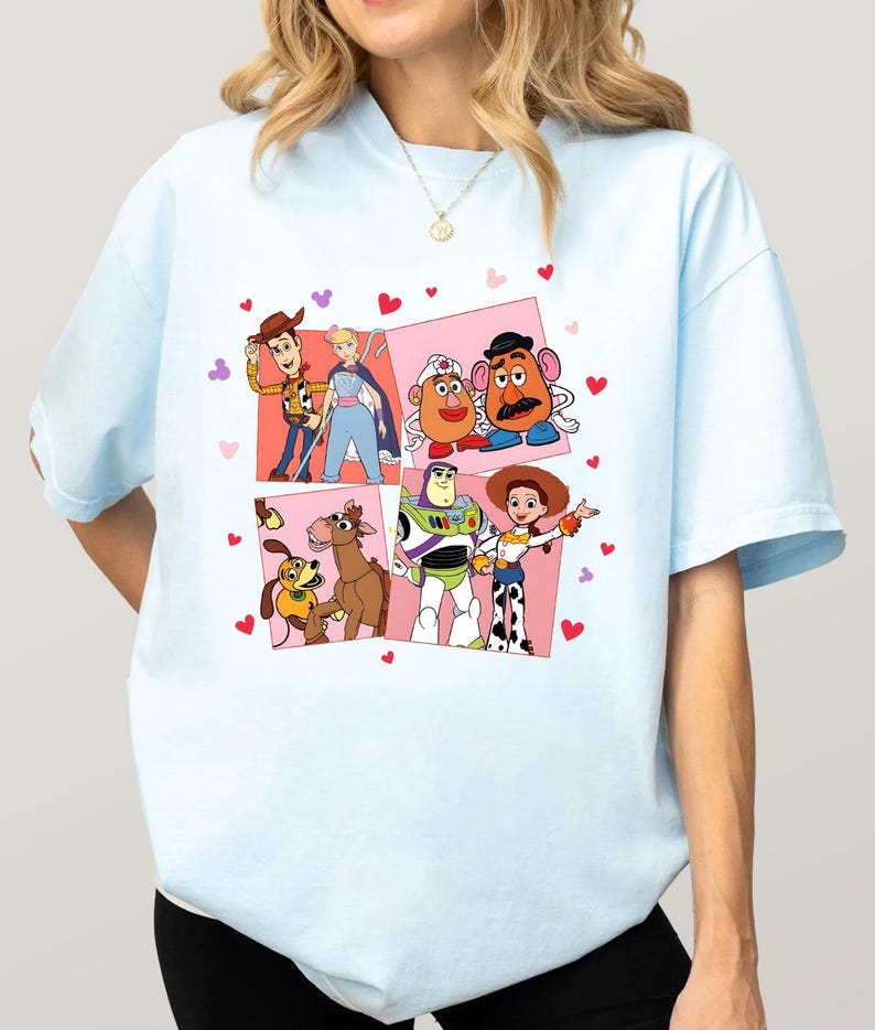 May include: Pale blue t-shirt with a vibrant graphic of Toy Story characters. The design showcases Woody, Buzz Lightyear, Jessie, and others, set against pink squares and red heart motifs.