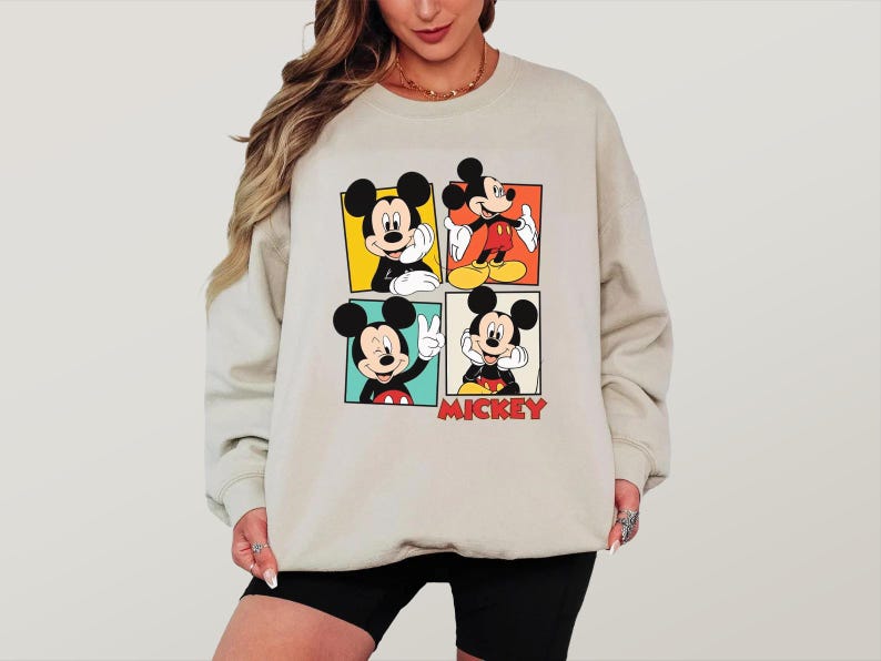 Peut inclure: Un sweat-shirt beige avec quatre panneaux repr&eacute;sentant diff&eacute;rentes images de Mickey Mouse. Chaque panneau montre Mickey dans une pose diff&eacute;rente, avec le mot "MICKEY" en lettres rouges en bas &agrave; droite.