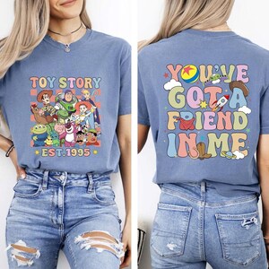 May include: A light blue t-shirt featuring a colorful graphic of Toy Story characters with the text "TOY STORY EST.1995" on the front. The back of the shirt displays the phrase "YOU'VE GOT A FRIEND IN ME" with related graphics.