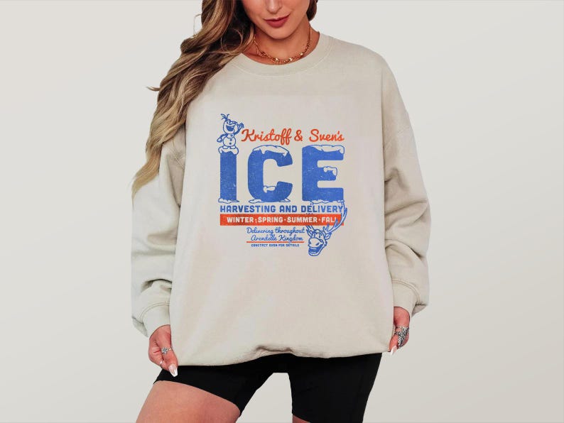 May include: A cream-coloured sweatshirt featuring a blue and red graphic that says "Kristoff & Sven's ICE Harvesting and Delivery." The design includes a snowman and reindeer illustration. The sweatshirt is long-sleeved with a crew neck.