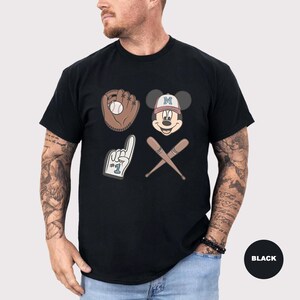 May include: Black t-shirt featuring baseball-themed graphics. The design includes a baseball glove with a ball, Mickey Mouse wearing a baseball cap, a foam finger, and crossed baseball bats. The baseball cap has the letter "M" on it.