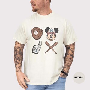 May include: Cream-colored t-shirt featuring baseball-themed graphics. The design includes a baseball glove with a ball, Mickey Mouse wearing a baseball cap, a foam finger, and crossed baseball bats. The word "NATURAL" is printed on the bottom right.