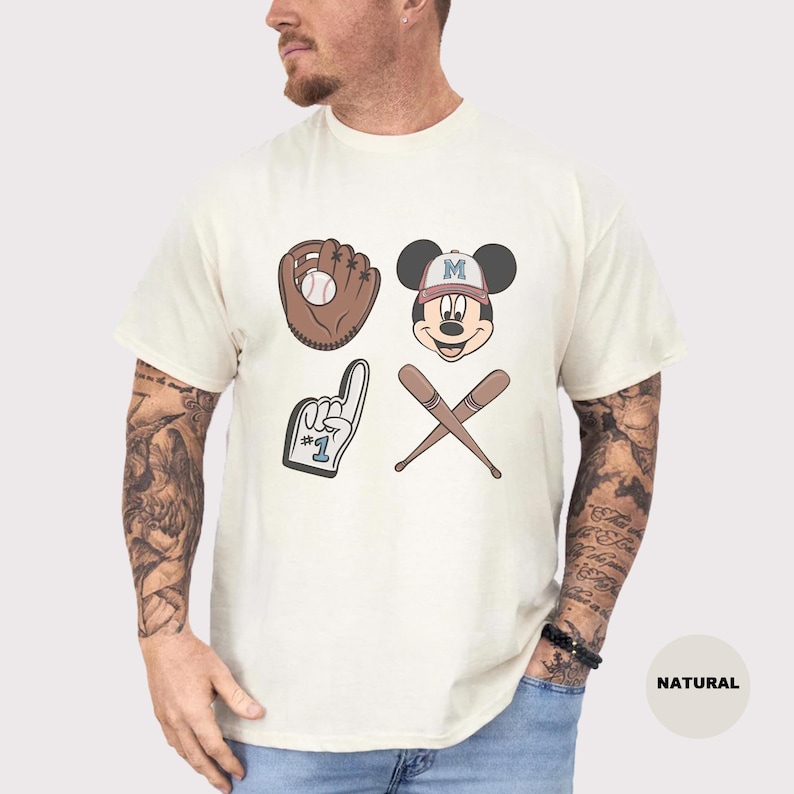 May include: Cream-colored t-shirt featuring baseball-themed graphics. The design includes a baseball glove with a ball, Mickey Mouse wearing a baseball cap, a foam finger, and crossed baseball bats. The word "NATURAL" is printed on the bottom right.