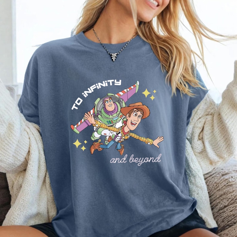 May include: Blue t-shirt with a graphic of Buzz Lightyear and Woody from Toy Story, and the text "TO INFINITY and beyond". The design includes yellow stars and a colourful spaceship wing.