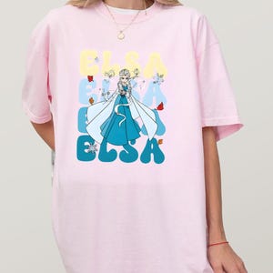 May include: Light pink oversized t-shirt featuring a graphic of Elsa from Frozen. The design includes the name "ELSA" in a retro font, with Elsa in a blue dress and white cape. Butterflies and snowflakes are also included in the design.