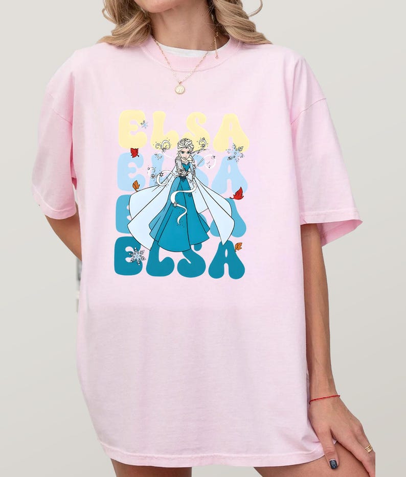 May include: Light pink oversized t-shirt featuring a graphic of Elsa from Frozen. The design includes the name "ELSA" in a retro font, with Elsa in a blue dress and white cape. Butterflies and snowflakes are also included in the design.