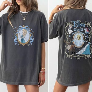 May include: Dark gray t-shirt featuring a front graphic of a princess in a blue dress within a gold frame and blue ribbon. The back of the shirt displays the name "Elsa" and additional character illustrations in a decorative frame. The shirt is a casual, oversized fit.