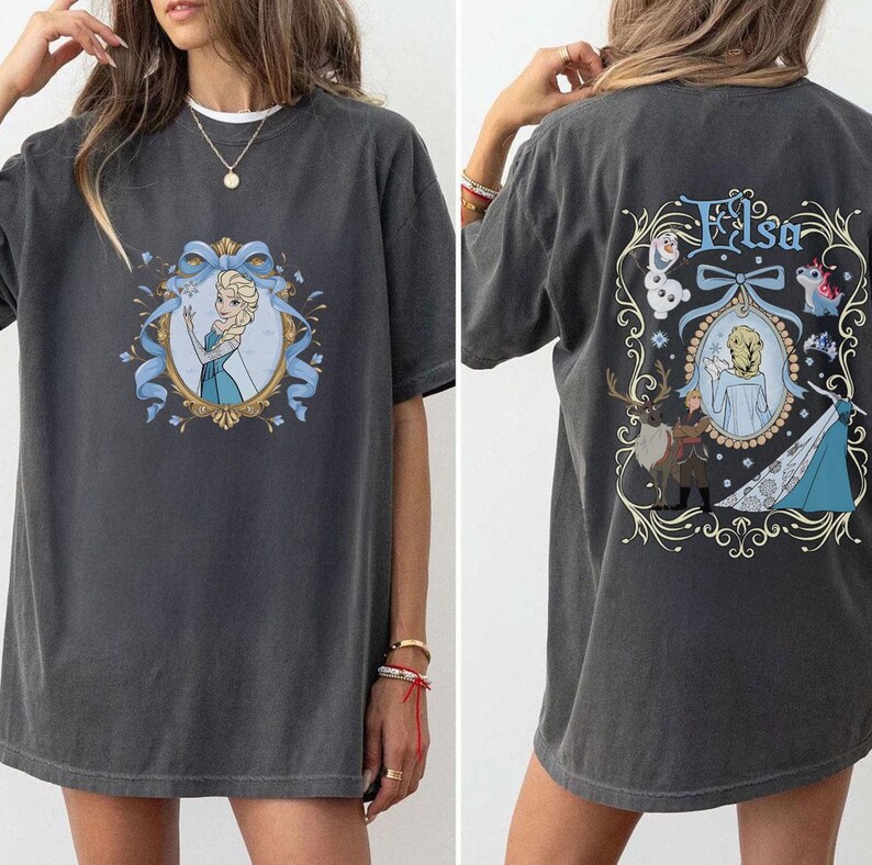 May include: Dark gray t-shirt featuring a front graphic of a princess in a blue dress within a gold frame and blue ribbon. The back of the shirt displays the name "Elsa" and additional character illustrations in a decorative frame. The shirt is a casual, oversized fit.