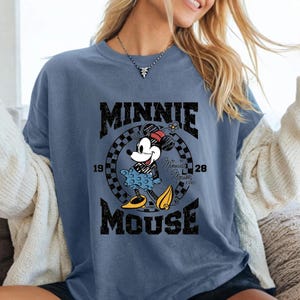 Może przedstawiać: Niebieska koszulka z grafiką Myszki Minnie. Projekt zawiera słowa "MINNIE MOUSE" w postarzonej czcionce, a także rok "1928". Myszka Minnie jest przedstawiona w swoim klasycznym stroju.