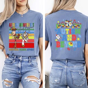 K&ouml;nnte beinhalten: Hellblaues T-Shirt mit der Aufschrift "AUTISM AWARENESS And Beyond" und den Zeichentrickfiguren Woody und Buzz Lightyear. Auf der R&uuml;ckseite des Shirts steht "TO AUTISM AND BEYOND" mit einer Rakete und Puzzleteilen.
