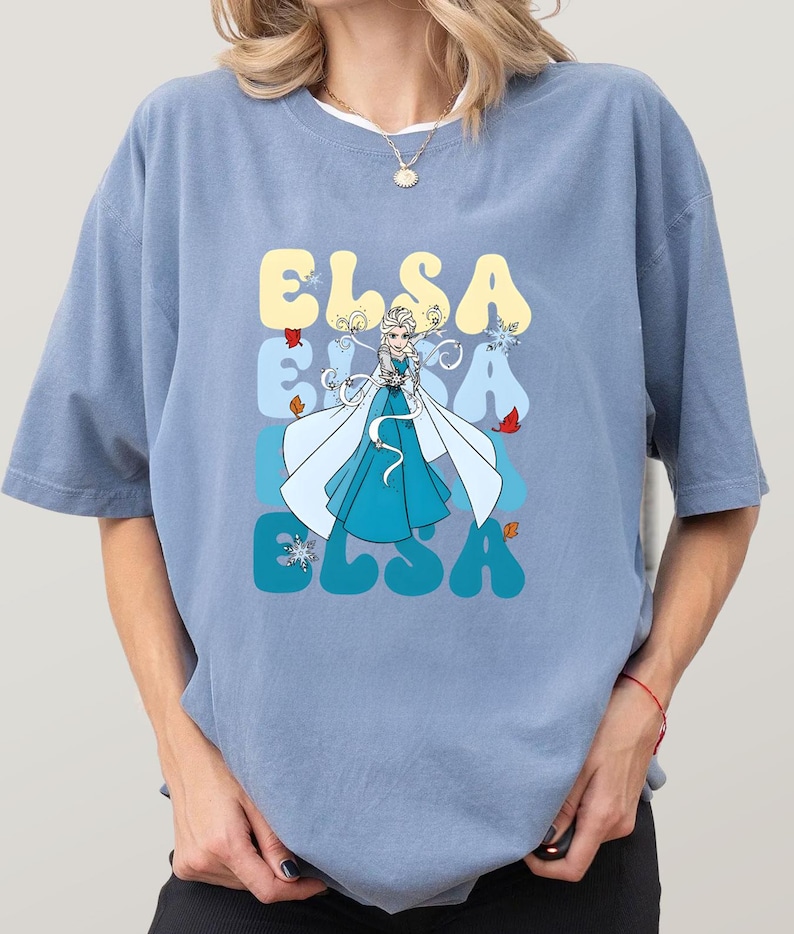 May include: A light blue t-shirt featuring a graphic of Elsa from Frozen. The word "ELSA" is stacked vertically in yellow, blue, and teal. Elsa is depicted in a blue dress and white cape, with snowflakes and leaves.