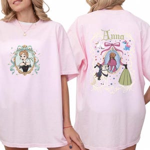 May include: Pink t-shirt featuring a cartoon image of a woman in a black dress within a decorative frame on the front. The back of the shirt has a matching design with the name "Anna" and a mirror image.
