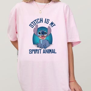 Puede incluir: Camiseta rosa claro con una imagen de dibujos animados de Stitch de Lilo & Stitch. El personaje azul de Stitch sonr&iacute;e, con el texto "Stitch is my Spirit Animal" en letras azules. La camiseta es de estilo casual y oversize.