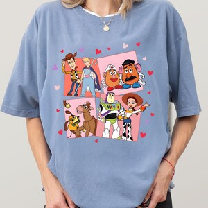 May include: A pale blue t-shirt with a colourful graphic of Toy Story characters. The design features Woody, Buzz Lightyear, Jessie, and other characters in a pink square with hearts.