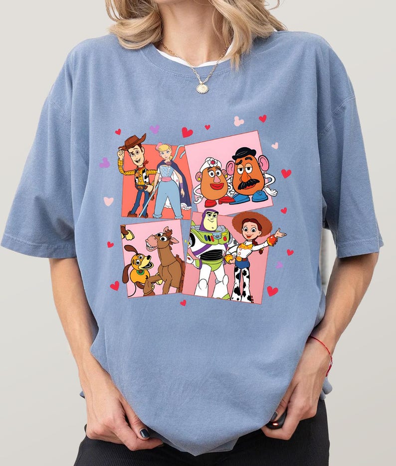 May include: A pale blue t-shirt with a colourful graphic of Toy Story characters. The design features Woody, Buzz Lightyear, Jessie, and other characters in a pink square with hearts.