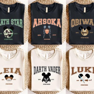 Op de afbeelding: Zes T-shirts met Star Wars-ontwerpen. De shirts tonen de namen "Death Star", "Ahsoka", "Obiwan", "Leia", "Darth Vader" en "Luke" in een collegiale lettertype, samen met het jaar "1977" en het Star Wars-logo. Elk ontwerp bevat een Mickey Mouse silhouet.