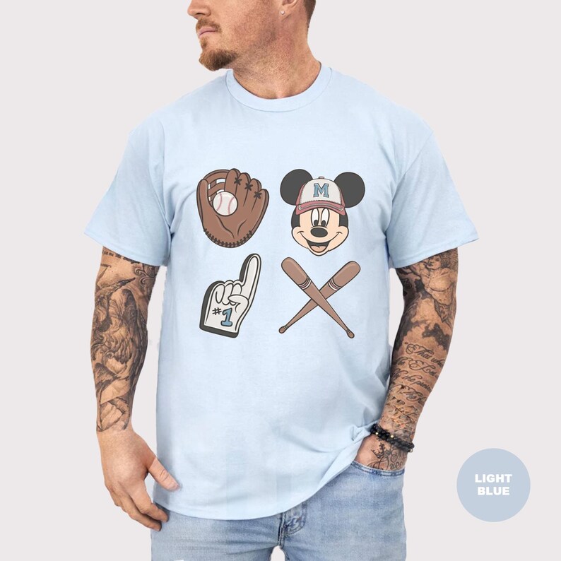May include: Light blue t-shirt featuring baseball-themed graphics. The design includes a baseball glove with a ball, Mickey Mouse with a baseball cap, crossed baseball bats, and a foam finger. The shirt is a light blue color.