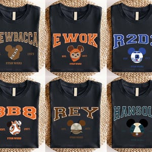 Op de afbeelding: Zes zwarte t-shirts met Star Wars-personagenamen in collegiale letters. Elk shirt heeft de naam van een personage, een op Mickey Mouse ge&iuml;nspireerde afbeelding en de tekst "EST. 1977 STAR WARS". Personages zijn onder meer Chewbacca, Ewok, R2D2, BB8, Rey en Han Solo.
