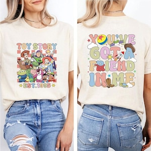 May include: Beige t-shirt featuring a colorful graphic of Toy Story characters with the text "TOY STORY EST.1995" on the front. The back of the shirt displays the phrase "YOU'VE GOT A FRIEND IN ME" with various Toy Story themed illustrations.