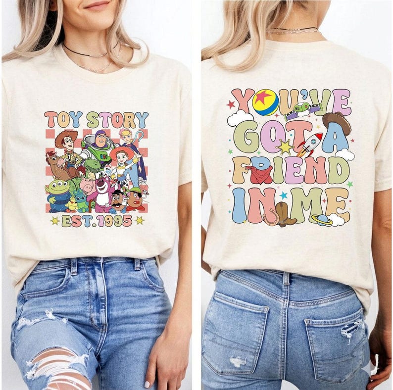 May include: Beige t-shirt featuring a colorful graphic of Toy Story characters with the text "TOY STORY EST.1995" on the front. The back of the shirt displays the phrase "YOU'VE GOT A FRIEND IN ME" with various Toy Story themed illustrations.