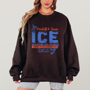 May include: A dark brown sweatshirt with the text "Kristoff & Sven's ICE" in orange and blue. The sweatshirt also features the text "HARVESTING AND DELIVERY" and "WINTER-SPRING-SUMMER-FALL".