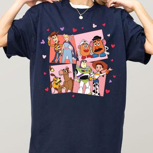 May include: Navy blue t-shirt with a colourful graphic of Toy Story characters. The design includes Woody, Buzz Lightyear, and other characters in a square pattern with heart and Mickey Mouse head accents. A white crew neck is visible.
