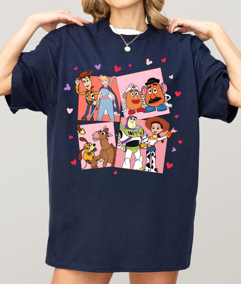 May include: Navy blue t-shirt with a colourful graphic of Toy Story characters. The design includes Woody, Buzz Lightyear, and other characters in a square pattern with heart and Mickey Mouse head accents. A white crew neck is visible.