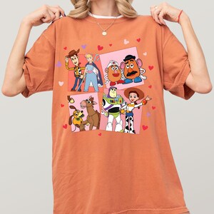 May include: An orange t-shirt with a colourful graphic of characters from the animated film Toy Story. The design includes images of Woody, Buzz Lightyear, and other characters, with pink squares and heart accents.