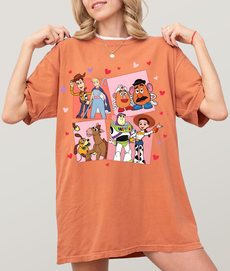 May include: An orange t-shirt with a colourful graphic of characters from the animated film Toy Story. The design includes images of Woody, Buzz Lightyear, and other characters, with pink squares and heart accents.