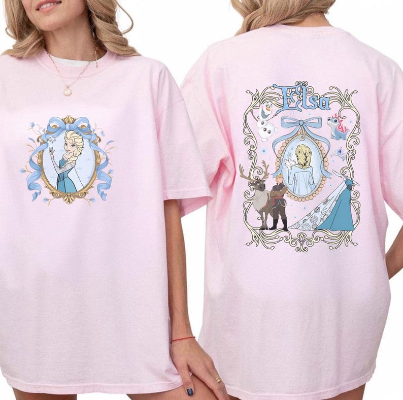 May include: Pink t-shirts featuring Elsa from Frozen. The front design shows Elsa in a blue dress within a decorative frame. The back of the shirt has the name "Elsa" and other characters from the movie.