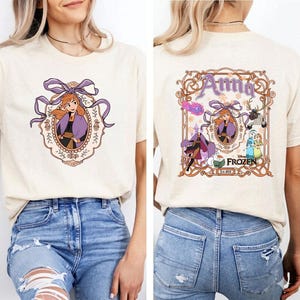 以下が含まれることがあります： ディズニーの「アナと雪の女王」のアナの漫画イラストが描かれたベージュのTシャツ。前面のデザインは、紫色のフレームとリボンでアナを描いています。シャツの背面には、アナと「アナ」の文字が入った大きなデザインがあります。