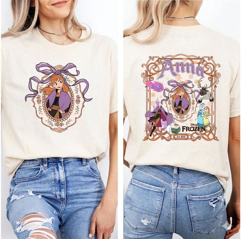 以下が含まれることがあります： ディズニーの「アナと雪の女王」のアナの漫画イラストが描かれたベージュのTシャツ。前面のデザインは、紫色のフレームとリボンでアナを描いています。シャツの背面には、アナと「アナ」の文字が入った大きなデザインがあります。