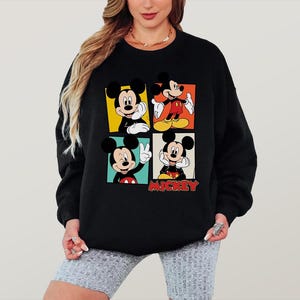 Peut inclure: Sweat-shirt noir avec quatre panneaux repr&eacute;sentant Mickey Mouse dans diff&eacute;rentes poses. Chaque panneau a un fond de couleur diff&eacute;rente : jaune, orange, rouge et vert. Le mot "Mickey" est en lettres rouges en bas.