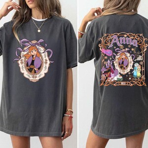 以下が含まれることがあります： 紫と黒の衣装を着たキャラクターの漫画のイメージが特徴の濃い灰色のTシャツ。背中には「Anna」という名前が書かれています。前面のデザインは紫のリボンが付いた楕円形のフレームに入っています。背面のデザインには他のキャラクターが含まれています。