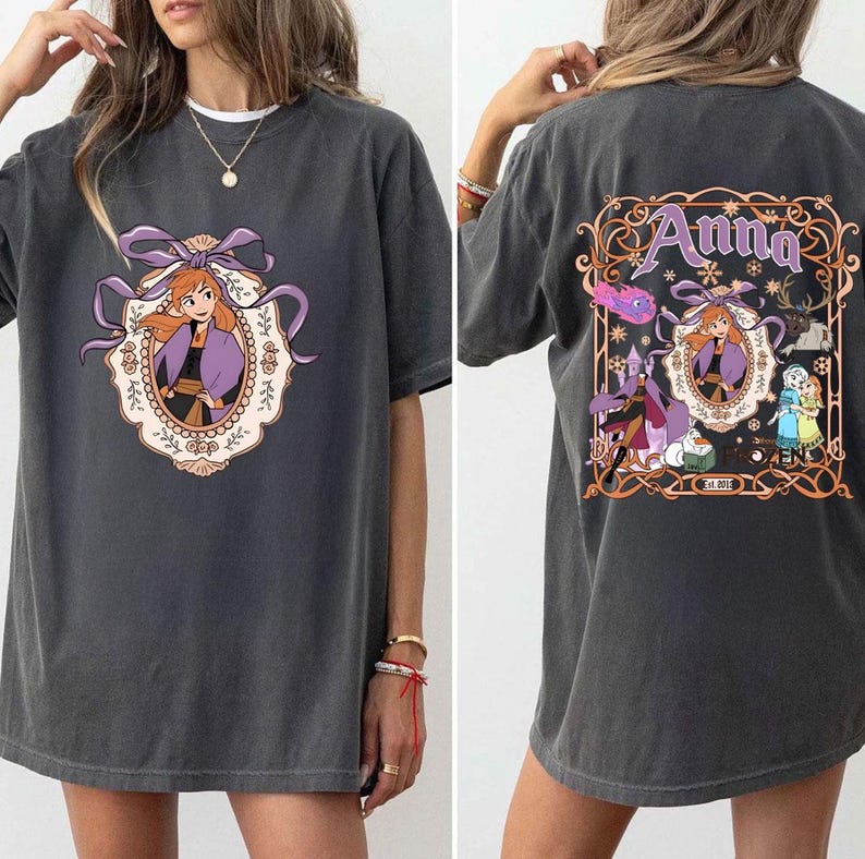 以下が含まれることがあります： 紫と黒の衣装を着たキャラクターの漫画のイメージが特徴の濃い灰色のTシャツ。背中には「Anna」という名前が書かれています。前面のデザインは紫のリボンが付いた楕円形のフレームに入っています。背面のデザインには他のキャラクターが含まれています。