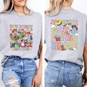 May include: A light gray t-shirt featuring a colorful graphic of Toy Story characters with the text "TOY STORY EST.1995" on the front. The back of the shirt displays the phrase "YOU'VE GOT A FRIEND IN ME" with related graphics.