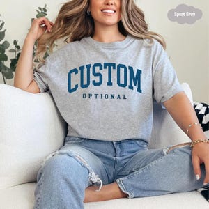 Peut inclure: Un t-shirt gris clair à manches courtes avec le mot "CUSTOM" en lettres majuscules bleu foncé au-dessus du mot "OPTIONAL". Le t-shirt est fait d'une matière douce.
