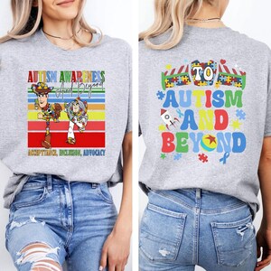 K&ouml;nnte beinhalten: Ein hellgraues T-Shirt mit einer farbenfrohen Grafik zur F&ouml;rderung des Autismus-Bewusstseins. Auf der Vorderseite sind Woody und Buzz Lightyear mit dem Text "Autism Awareness and Beyond" abgebildet. Auf der R&uuml;ckseite steht "To Autism and Beyond" mit Puzzleteilen und einer Rakete.