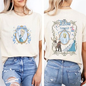 May include: Beige t-shirt featuring a cartoon image of a princess in a blue dress, surrounded by a blue ribbon and floral design. The back of the shirt has the word "Elsa" and other cartoon characters in a decorative frame.