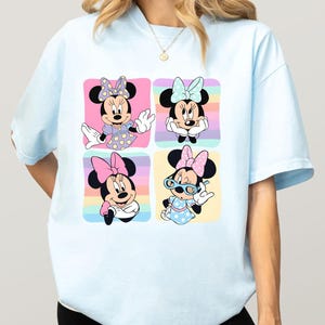 Może przedstawiać: Jasnoniebieska koszulka z czterema panelami przedstawiającymi Minnie Mouse w r&oacute;żnych pozach. Każdy panel ma pastelowe tęczowe tło. Minnie ma na sobie r&oacute;żne stroje i kokardy w każdym panelu.