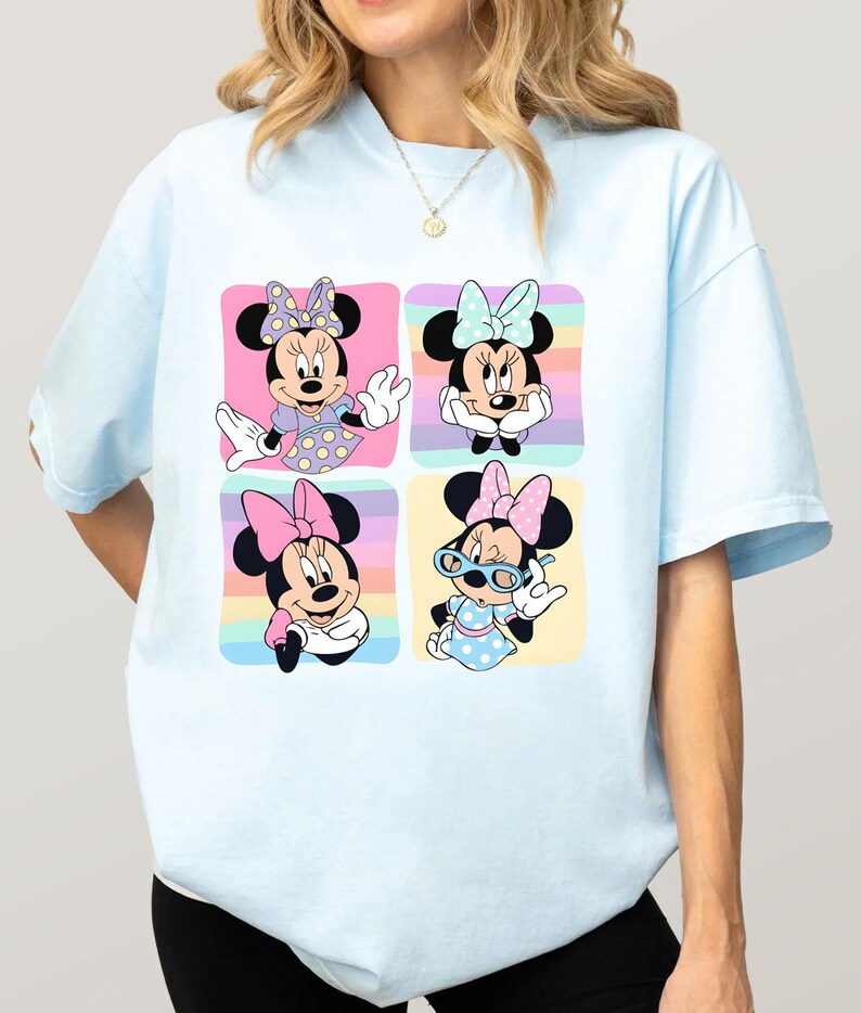 Może przedstawiać: Jasnoniebieska koszulka z czterema panelami przedstawiającymi Minnie Mouse w r&oacute;żnych pozach. Każdy panel ma pastelowe tęczowe tło. Minnie ma na sobie r&oacute;żne stroje i kokardy w każdym panelu.