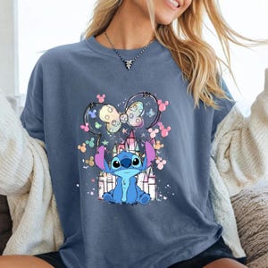 May include: A blue t-shirt with a cartoon image of Stitch from Lilo & Stitch. The design includes a castle, a bow, and colorful Mickey Mouse silhouettes. The shirt is a solid blue colour.