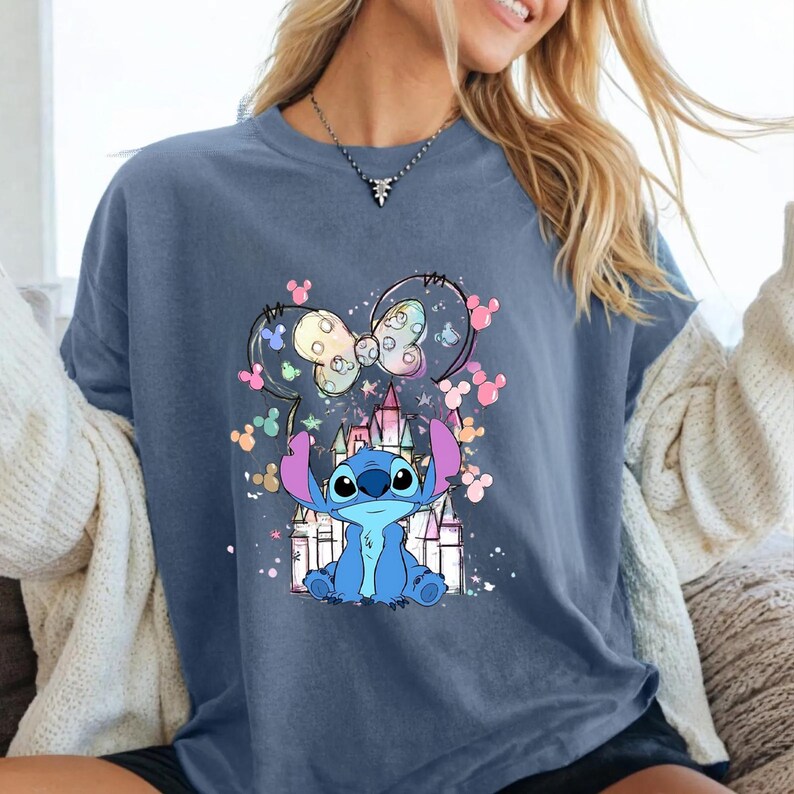 May include: A blue t-shirt with a cartoon image of Stitch from Lilo & Stitch. The design includes a castle, a bow, and colorful Mickey Mouse silhouettes. The shirt is a solid blue colour.