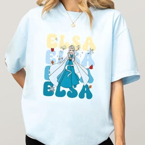 May include: Light blue t-shirt featuring a cartoon illustration of Elsa from Frozen. The word "ELSA" is printed in a retro font in yellow and blue. The design includes snowflakes and autumn leaves.