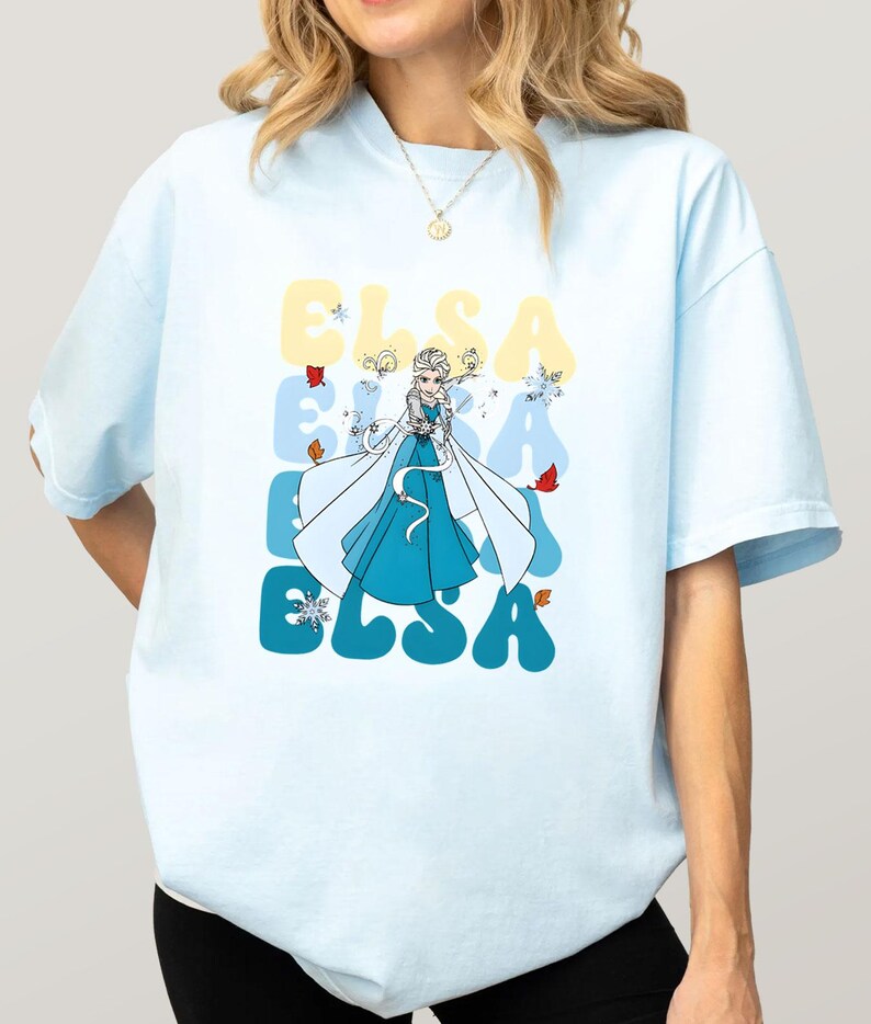 May include: Light blue t-shirt featuring a cartoon illustration of Elsa from Frozen. The word "ELSA" is printed in a retro font in yellow and blue. The design includes snowflakes and autumn leaves.