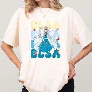 May include: Cream-colored t-shirt featuring a cartoon illustration of Elsa from Frozen. The word "ELSA" is printed in large, stylized letters in shades of blue and yellow. Elsa is depicted in her iconic blue dress.