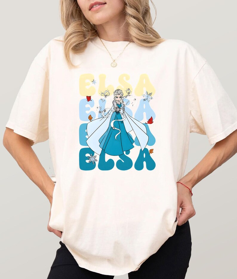 May include: Cream-colored t-shirt featuring a cartoon illustration of Elsa from Frozen. The word "ELSA" is printed in large, stylized letters in shades of blue and yellow. Elsa is depicted in her iconic blue dress.