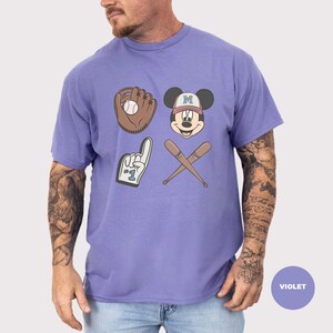 May include: Violet t-shirt featuring baseball-themed graphics. The design includes a baseball glove with a ball, Mickey Mouse's face wearing a baseball cap, a foam finger, and crossed baseball bats. The shirt is a solid violet color.