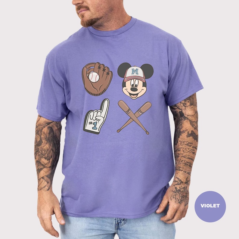 May include: Violet t-shirt featuring baseball-themed graphics. The design includes a baseball glove with a ball, Mickey Mouse's face wearing a baseball cap, a foam finger, and crossed baseball bats. The shirt is a solid violet color.